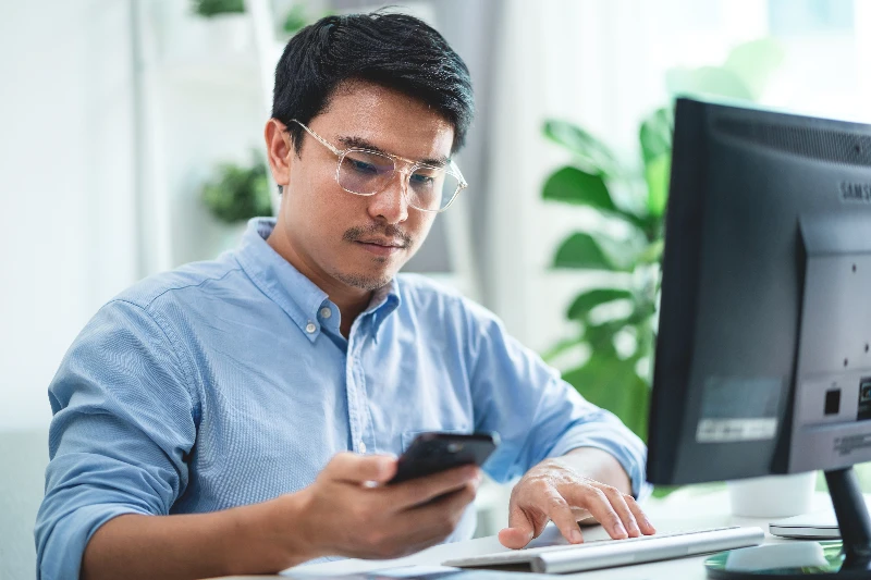 A man wearing glasses is sitting at a desk computer with a cell phone